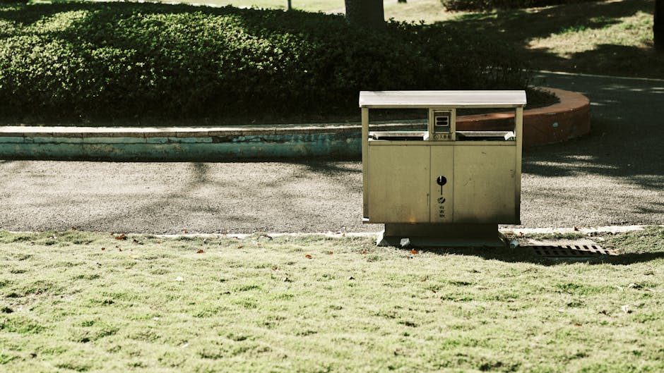 A small, metallic garden waste collection box with a lock and slot for depositing biodegradable garden debris, situated on a grassy verge beside a grey, textured asphalt pathway. Behind the box, there is a neatly trimmed hedge with dense green foliage and a curved, low brick border. The foreground features freshly mown grass with visible blade marks, and the environment is illuminated by soft, natural daylight, suggesting a calm, well-maintained outdoor space suitable for gardening and landscaping work, aligning with garden waste rules for garden maintenance as outlined on the gardenersroehampton.org.uk website.